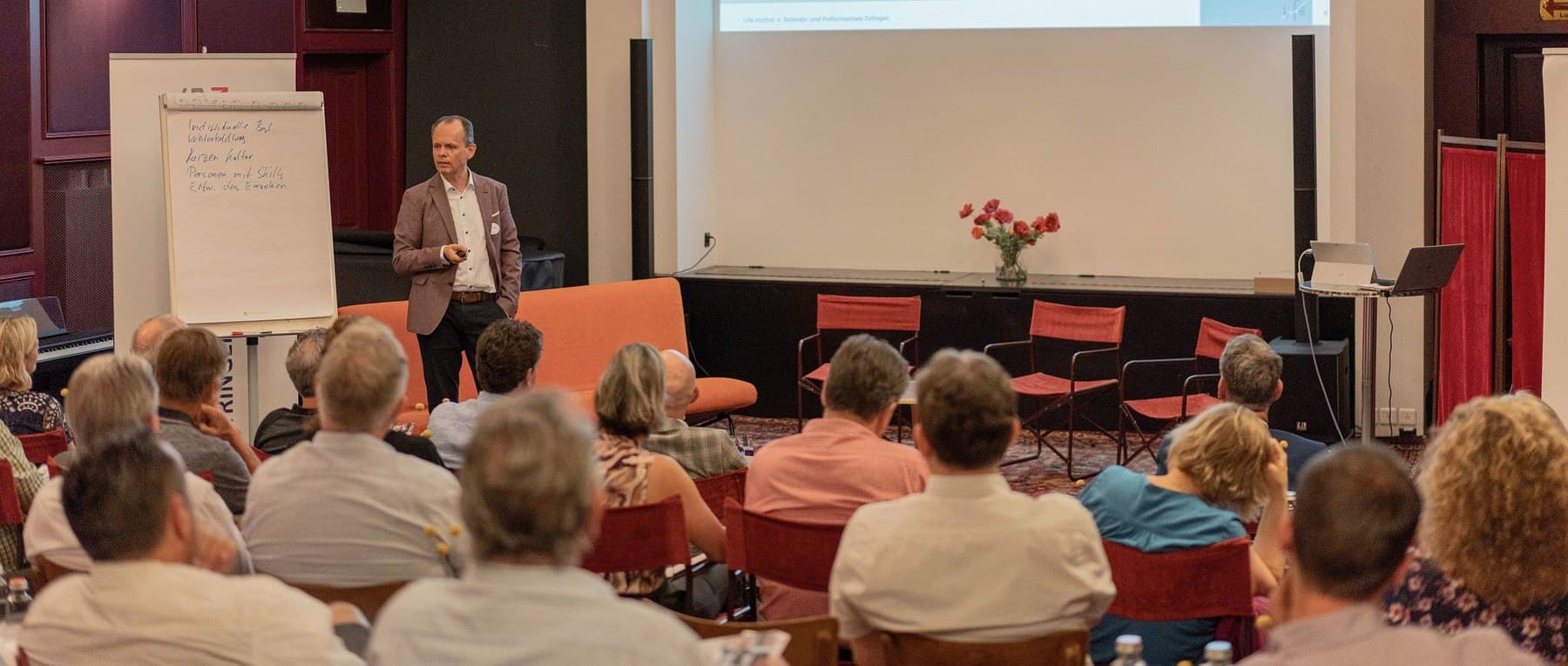 Hermann Ladner in front of a full hall at the Palass in Zofingen, the slide on screen reading Der explorative Dialog
